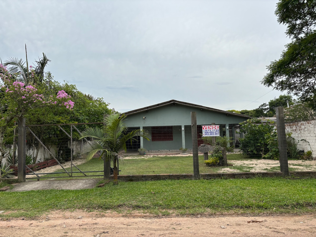 A single-story house is visible behind a fenced yard, featuring a manicured lawn and various plants, including pink flowering bushes. A 'For Sale' sign is displayed prominently on one side of the house, and pathways lead to the entrance.