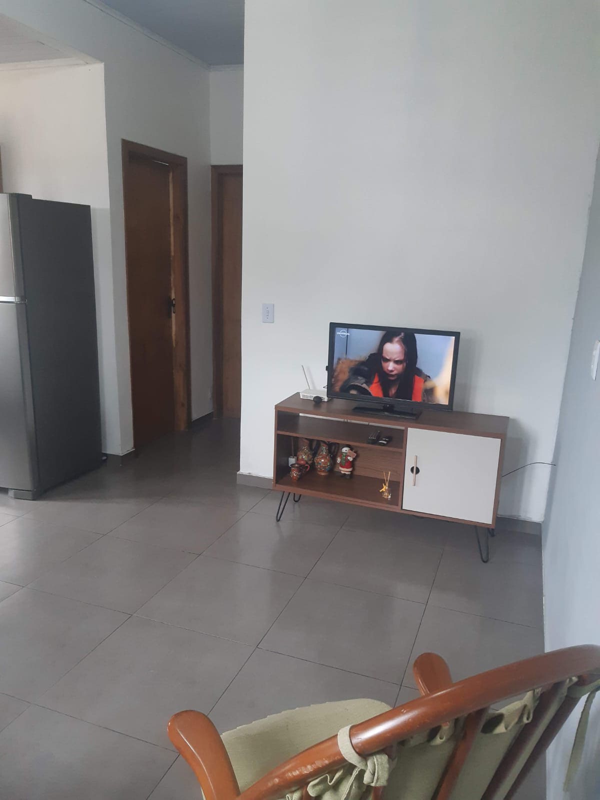 A living area is depicted with a television set positioned on a wooden TV stand, accompanied by decorative items. A chair is visible in the foreground, and doorways lead to other rooms. A refrigerator stands nearby, contributing to the functional layout.