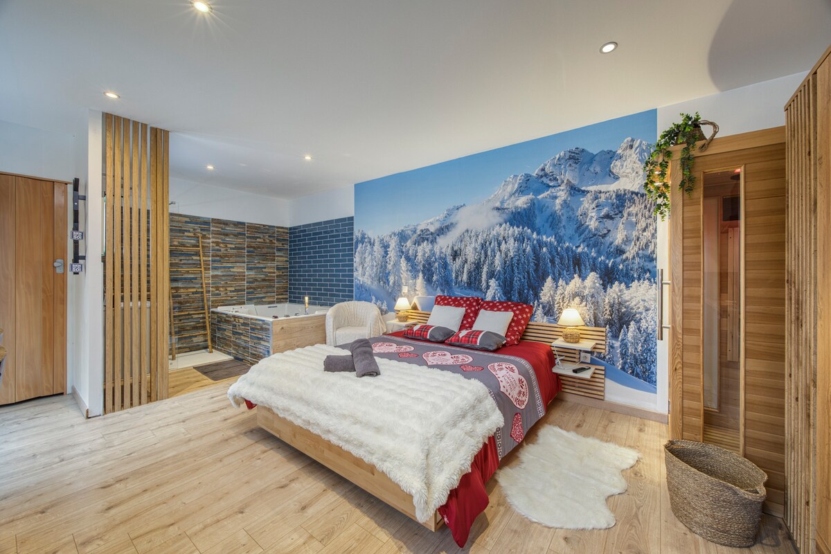 A spacious bedroom features a large bed topped with soft textiles, set against a scenic mural of snow-covered mountains. A cozy armchair, illuminated by bedside lamps, complements the natural wood accents throughout the room. An inviting bathroom area is visible, highlighting a relaxing bathtub.