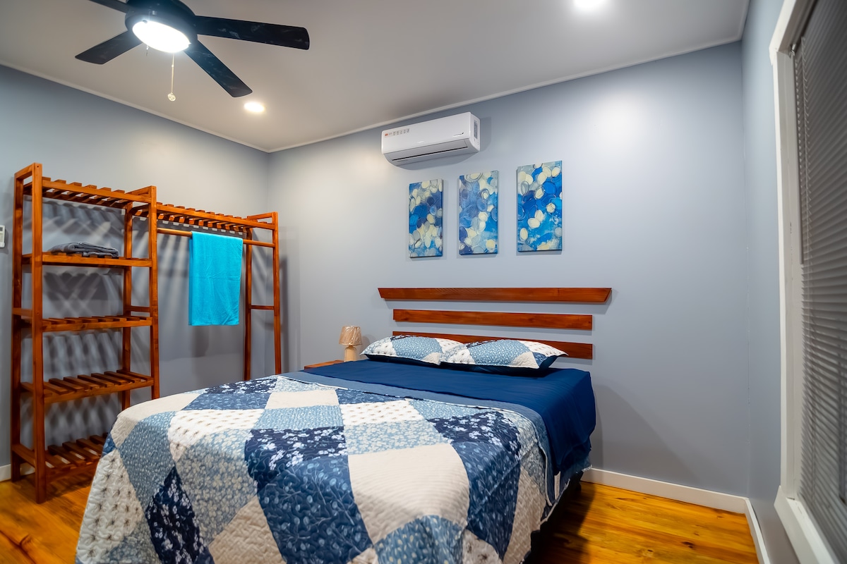 A cozy bedroom features a spacious bed adorned with a blue and white quilt. Above the bed, three pieces of wall art in shades of blue are displayed. A wooden rack holds blue towels, and a ceiling fan adds comfort. Natural light enters through a window with blinds.