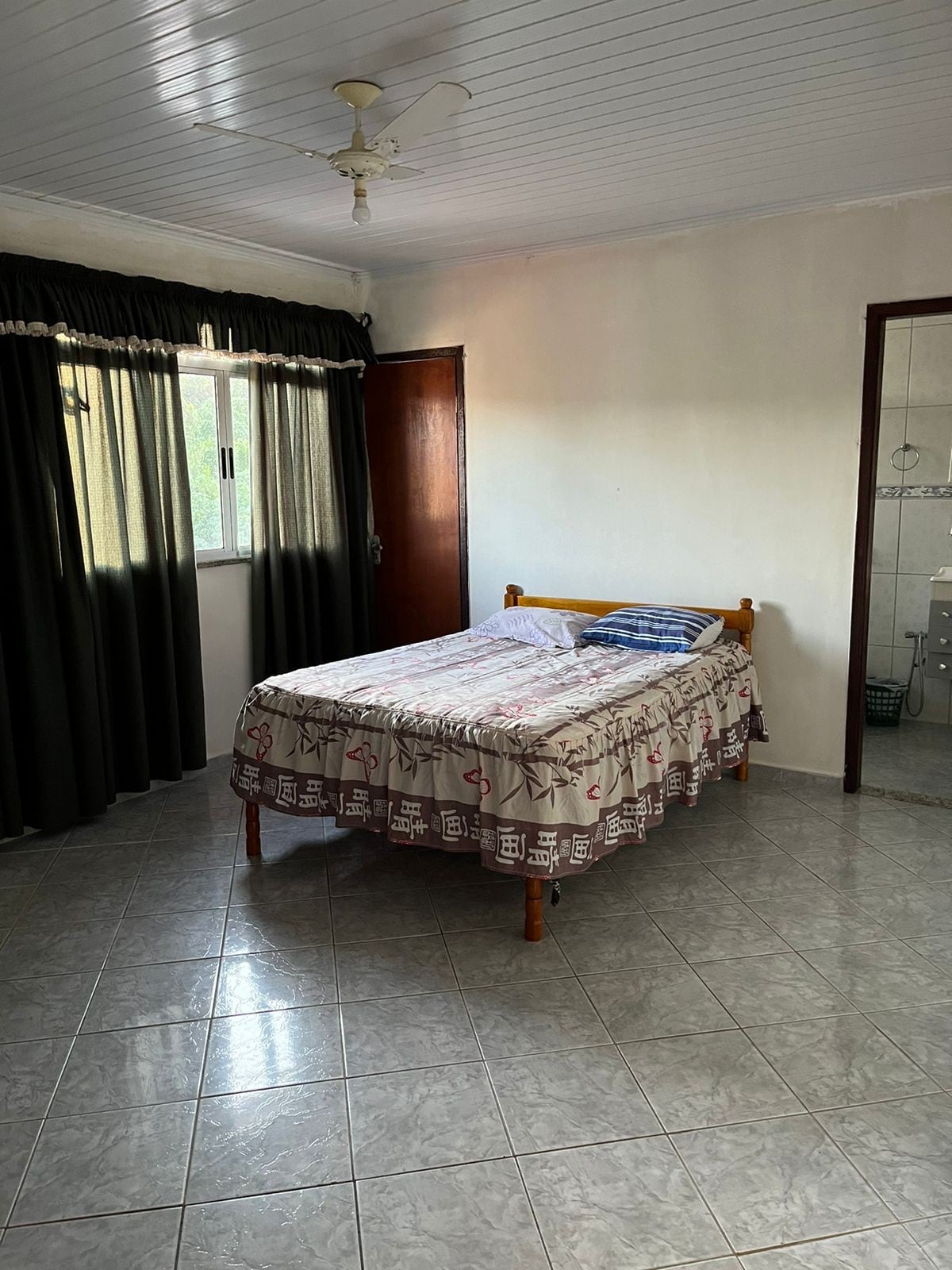 A simple bedroom features a wooden bed with a patterned blanket. Natural light enters through a window dressed with dark curtains. A ceiling fan is mounted above, and gray tile flooring contributes to a clean and functional design.