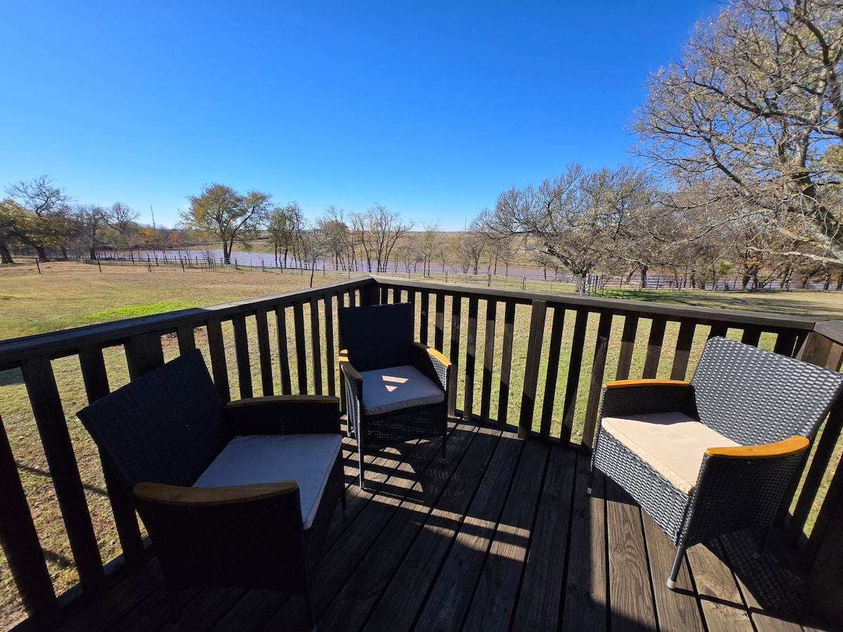 A private deck features two woven chairs with light cushions, surrounded by wooden railings. The expansive view reveals a serene landscape with a glimpse of a lake and trees in the distance under a clear blue sky.