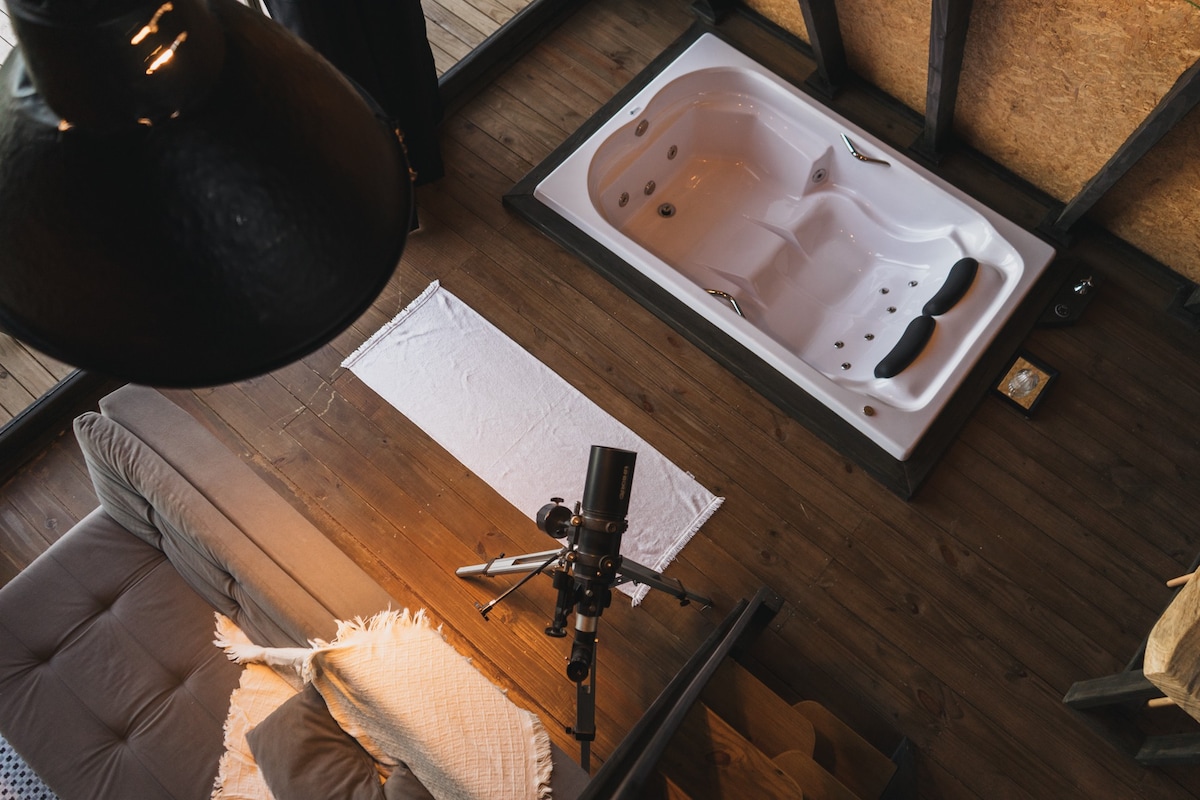 An aerial view captures a modern jacuzzi positioned within a wooden interior. Adjacent to the jacuzzi, a plush couch is partially visible, complemented by a soft blanket draped over it. A soft light illuminates the space, enhancing the relaxing atmosphere.