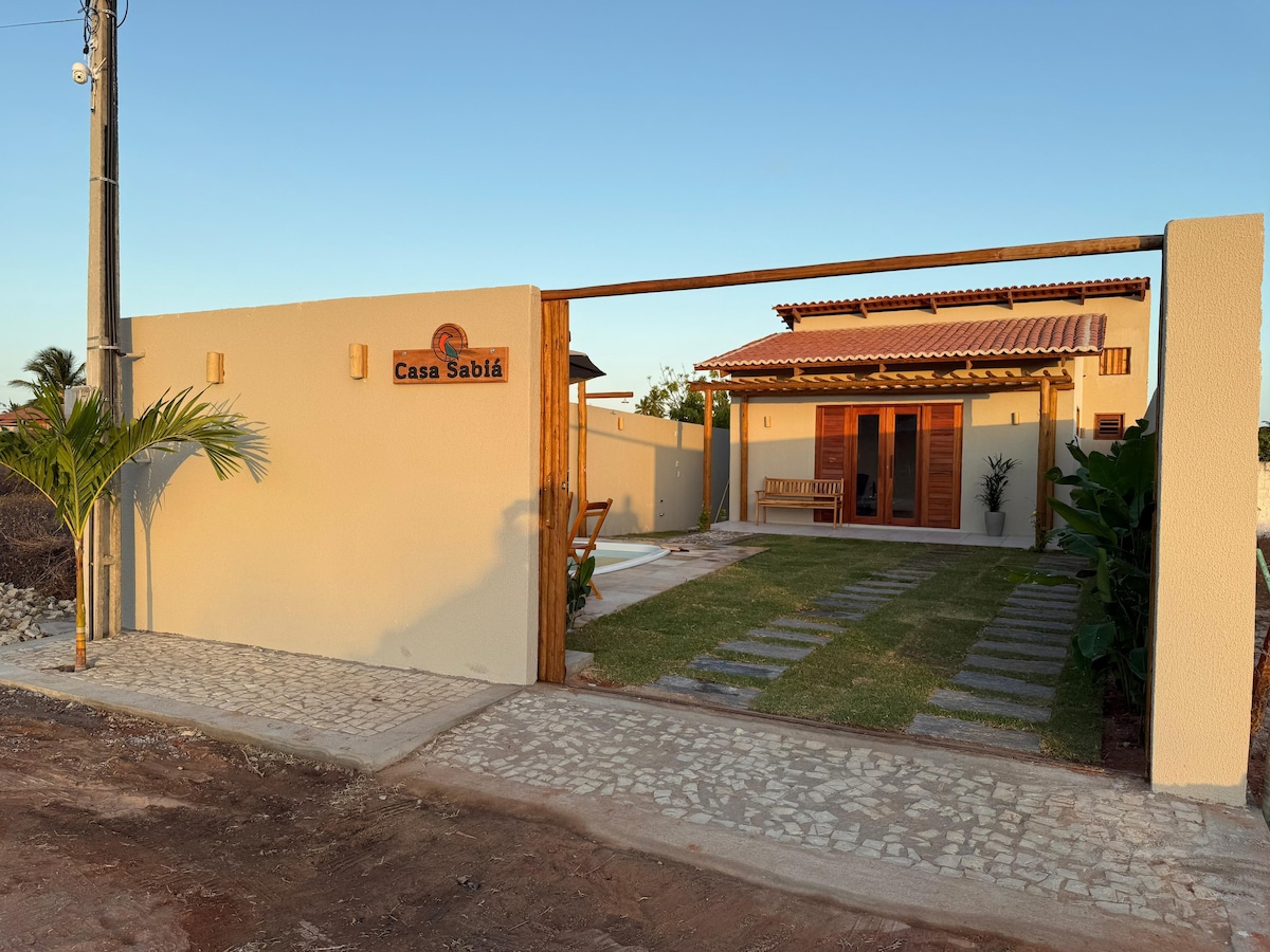 The exterior of the property is presented, featuring a welcoming entrance with a wooden gate, tan walls, and a small grassy area. A stone pathway leads through lush greenery, leading to a seating area and the house, which has a rustic tile roof.