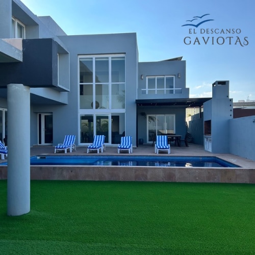 The exterior of a modern residence is featured, showcasing a private pool surrounded by lounge chairs with blue and white stripes. Extensive glass doors reflect the sky, while a vibrant green lawn provides a lush backdrop. The overall design emphasizes an inviting outdoor space.