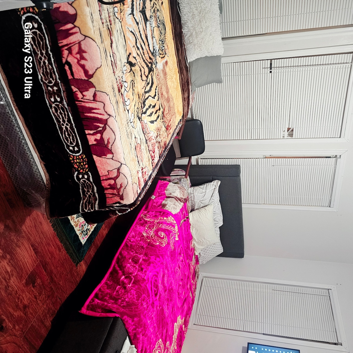 Two spacious queen beds are arranged in the room, each adorned with colorful blankets. A chair is positioned nearby. Large windows with closed shutters allow for natural light, and a Smart TV is visible in the corner, providing entertainment options.