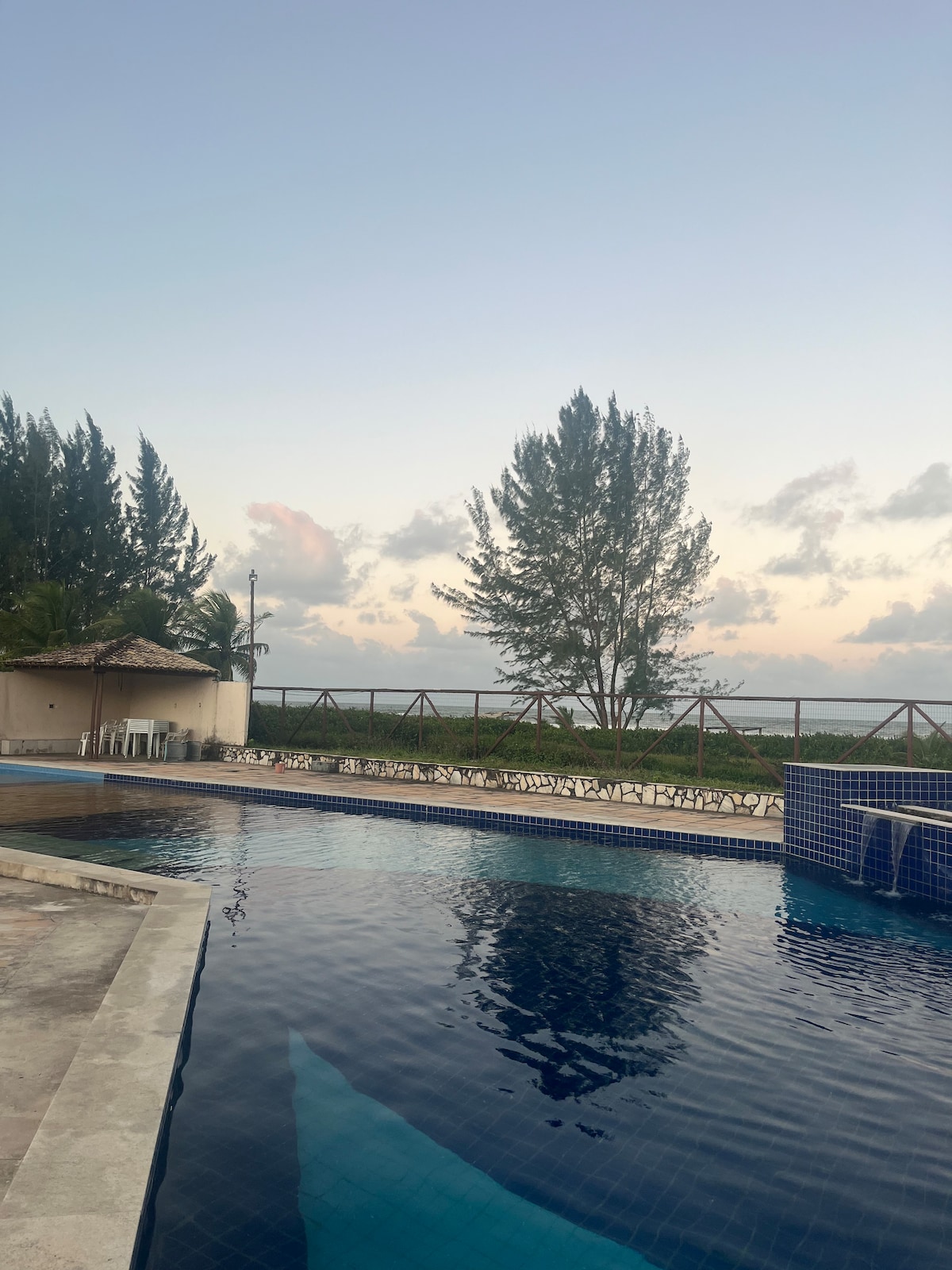 An outdoor pool reflects the soft colors of the sky during twilight. Lush greenery and tall trees border the pool area, while a gazebo provides shaded seating nearby. The tranquil water invites relaxation and bridges to the horizon.