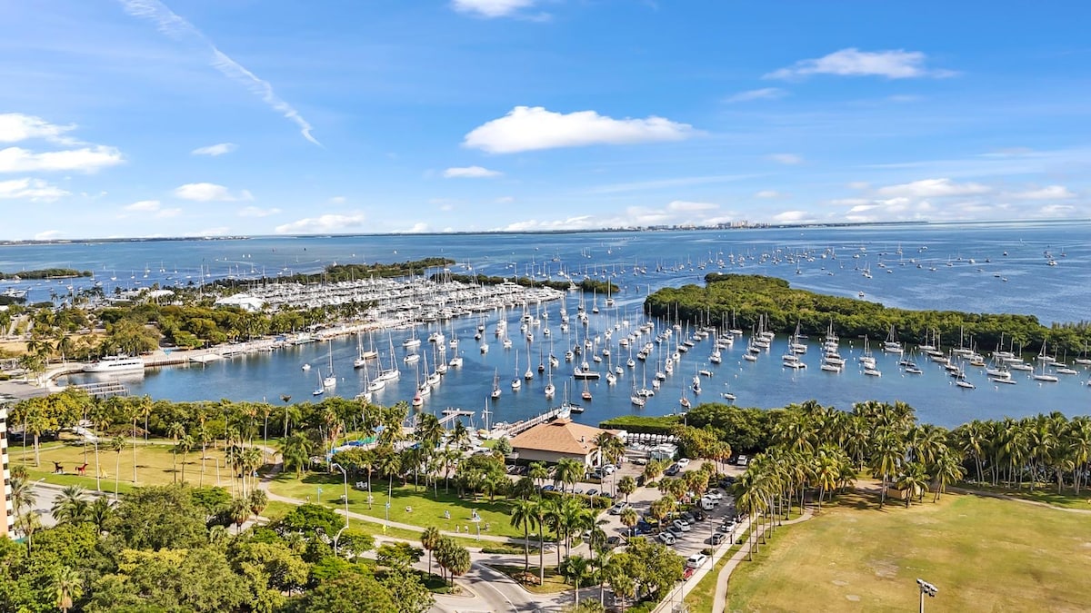 A panoramic view of a marina filled with numerous boats anchored in the water, surrounded by lush greenery and palm trees. An open area with grass is visible in the foreground, and a small building can be seen near the waterfront.