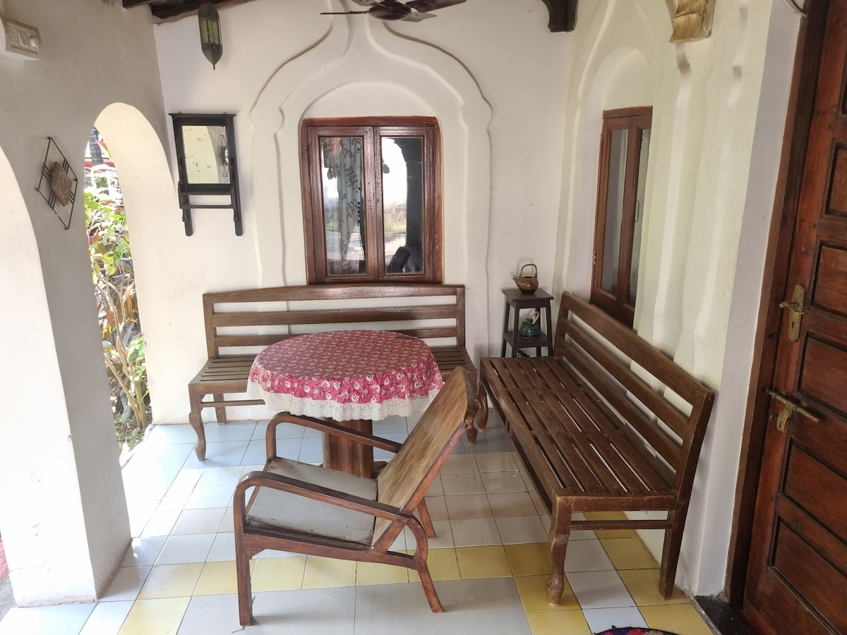 An outdoor seating area is featured, comprising a round table with a floral tablecloth and two wooden benches. A lounge chair is positioned nearby. White walls with wooden accents and a small window can be seen in the background, providing a sense of spaciousness.