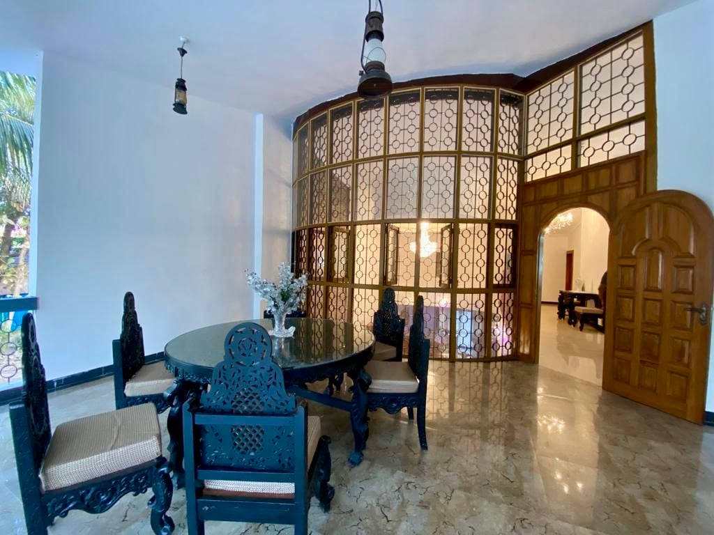 A dining area features a round black table surrounded by six intricately designed chairs. Large, decorative glass doors create a sense of openness, while a subtle floral arrangement sits at the center of the table. The polished marble floor adds an elegant touch.