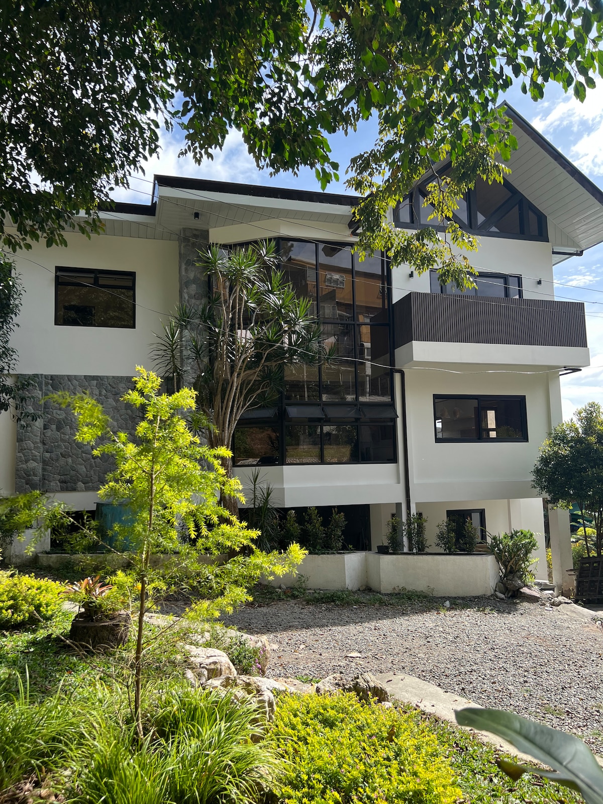 A modern three-story house is showcased surrounded by lush greenery. Large glass windows reflect the blue sky and sunlight while providing an open view of the outdoors. A landscaped garden with various plants and trees enhances the exterior, creating a serene environment.