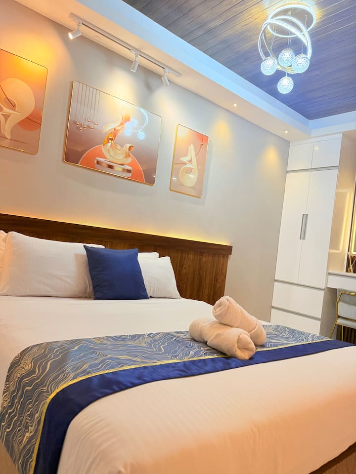 A stylish and restful bedroom featuring a comfy queen bed with crisp linens, plush towels, warm lighting, and modern décor. Includes a spacious white wardrobe and vanity area. A clean and inviting space perfect for a peaceful retreat.