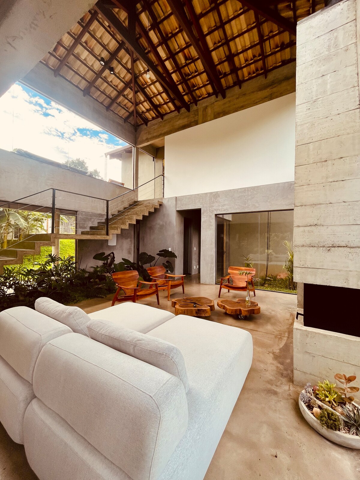 A spacious living area features a high, open ceiling with wooden beams and large windows that invite natural light. Cozy sofas are complemented by wooden chairs surrounding circular coffee tables. Indoor gardens add greenery to the tranquil environment.