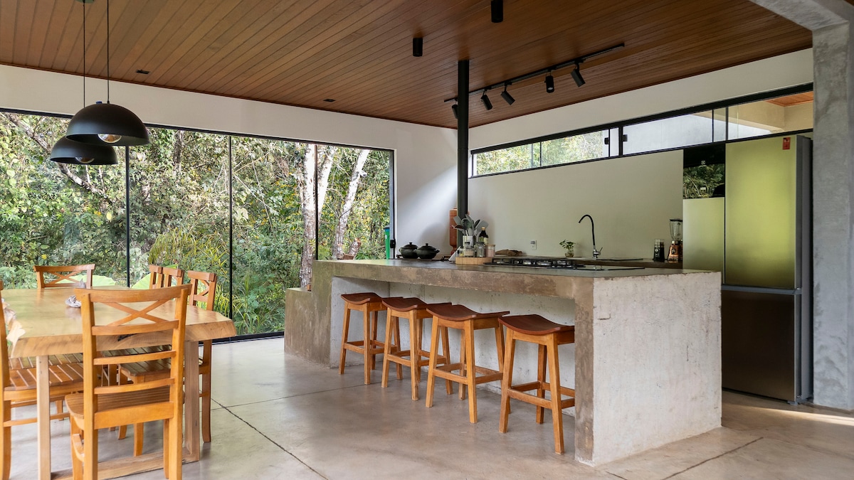 A spacious kitchen and dining area features large windows that frame views of the surrounding nature. The kitchen is equipped with modern appliances and rustic countertops, while the dining table, made of wood, seats eight and is complemented by wooden stools.