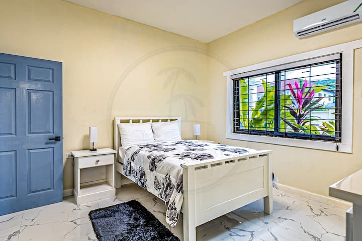 Táto svetlá, minimalistická spálňa má posteľ s kvetinovým vzorom, bielu komodu a nástenné zrkadlo. Oknom prúdi dovnútra prirodzené svetlo a odhaľuje bujnú zeleň vonku. Čisté línie a jemné tóny vytvárajú pokojné a príjemné útočisko.
