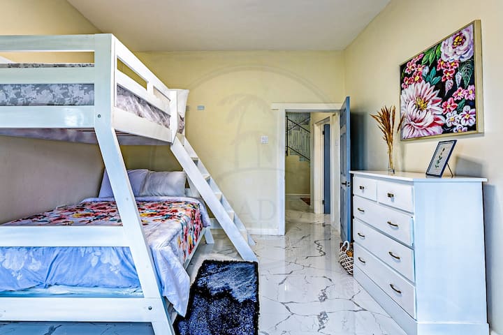 Kvetinová manželská posteľ a samostatná posteľ na poschodovej posteli, biela komoda a mäkké žlté steny. Čierny koberec pridáva kontrast a prirodzené svetlo z okna zvyšuje príjemnú atmosféru. Ideálne pre pohodlie a praktickosť v kompaktnom priestore.
