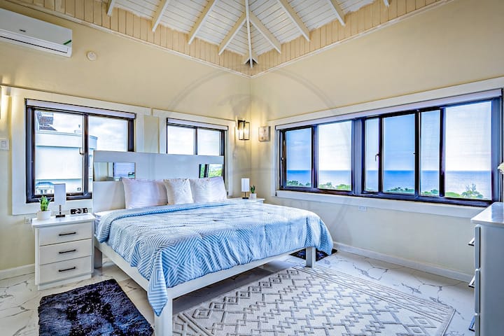 Táto príjemná hlavná spálňa má priestrannú posteľ s mäkkou modrou posteľnou bielizňou, teplé béžové steny a vysoké stropy s odhalenými trámami. Panoramatické okná zaplavujú miestnosť prirodzeným svetlom, zatiaľ čo vzorované koberce a lampy pri posteli dodávajú útulné čaro.
