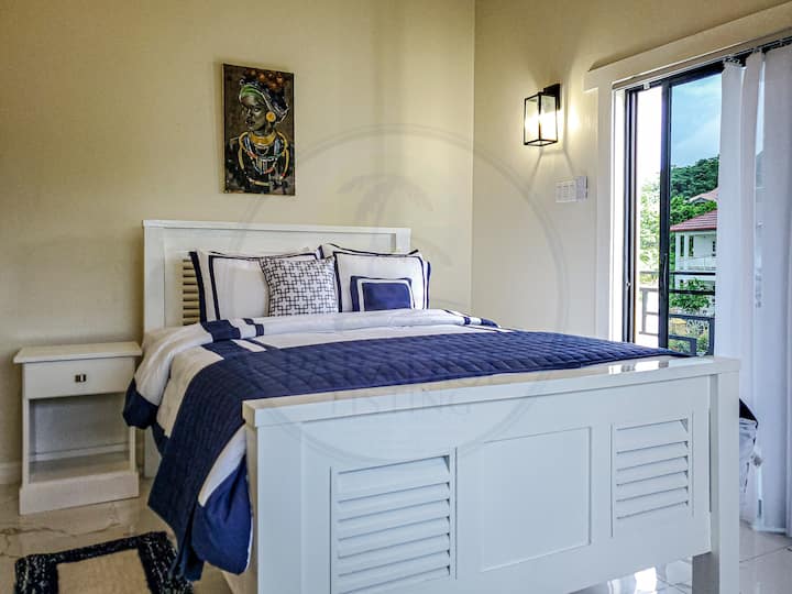 Táto útulná spálňa má veľkú posteľ s bielou posteľnou bielizňou a jemnou modrou dekou. Prirodzené svetlo preniká cez priesvitné závesy a zvýrazňuje odkryté trámy a krémové steny. Nástenná klimatizácia a bočný stolík dotvárajú pokojné prostredie.
