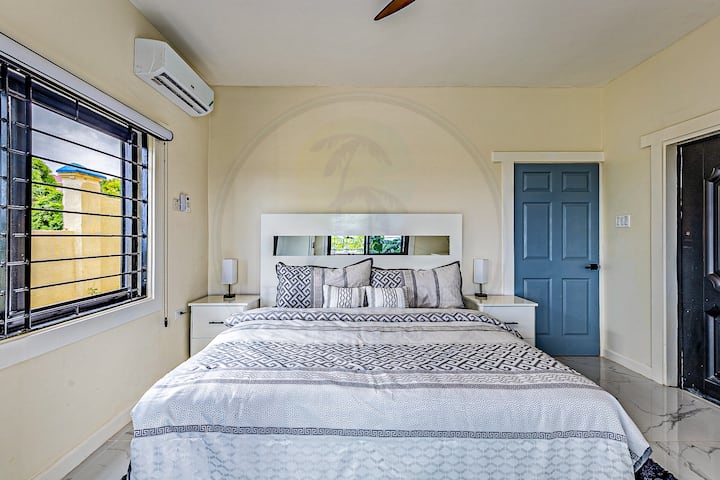 Táto minimalistická spálňa má veľkú posteľ s bielou a sivou posteľnou bielizňou, modré dvere a steny v jemných odtieňoch. Okno so žalúziami vpúšťa prirodzené svetlo, zatiaľ čo nástenná klimatizácia zaisťuje pohodlie. Priestor definujú čisté línie a jednoduchosť.
