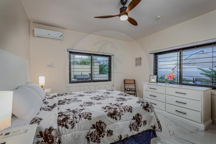 Táto svetlá spálňa má posteľnú prikrývku s kvetinovým vzorom, dva nočné stolíky s lampami a stropný ventilátor s drevenými lopatkami. Dve okná s čiernym rámom a rohová stolička dodávajú šarm, zatiaľ čo klimatizačná jednotka zaisťuje celoročné pohodlie.