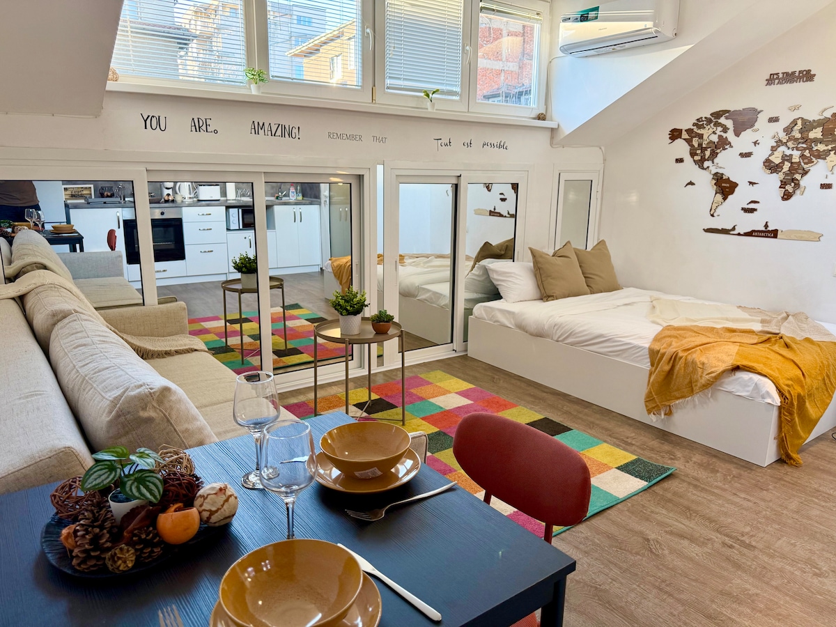 A well-lit studio showcases a cozy sleeping area with a spacious bed, complemented by a warm throw. The living space features a dining table set with dishes and a sofa, alongside mirrored cabinets that enhance the room's openness. A colorful area rug adds a welcoming touch.