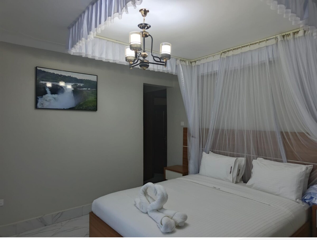 A spacious bedroom features a large bed with neatly arranged pillows. Light-colored bedding complements the serene atmosphere. A soft, decorative towel is shaped into an artistic design on the bed. A stylish chandelier hangs from the ceiling, casting a warm glow throughout the room.