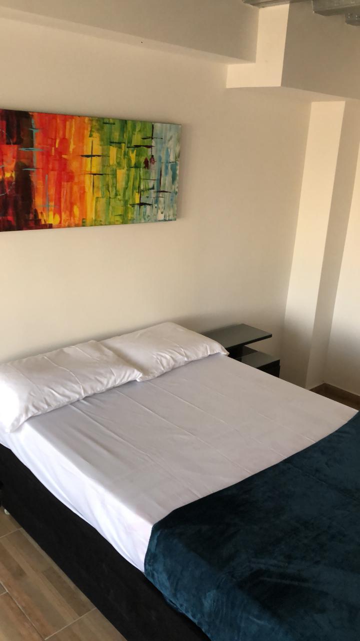 A well-lit sleeping area features a spacious bed with a white sheet and a dark green blanket. A bedside table stands beside the bed, and a colorful abstract artwork is displayed on the wall, adding a touch of vibrancy to the neutral-toned room.