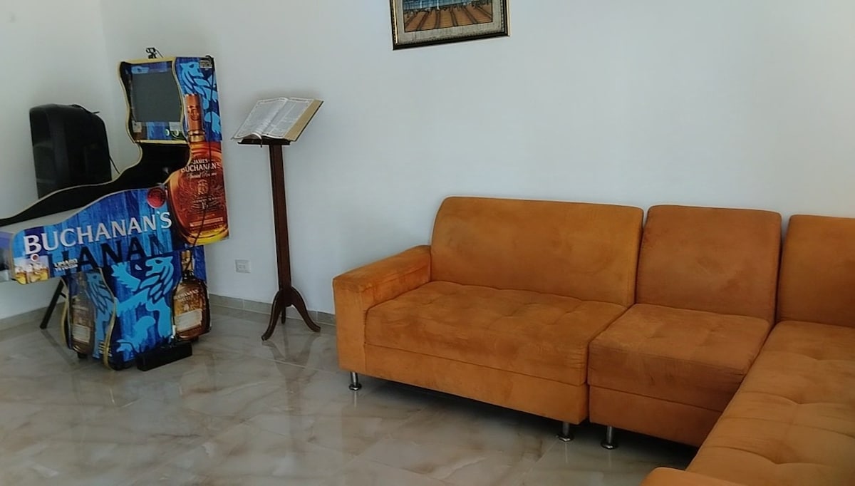 A spacious lounge area features a large orange sectional sofa arranged for comfort. An entertainment unit, designed with vibrant graphics, is positioned nearby, along with a stand displaying a music book. The marble flooring offers a polished appearance, enhancing the area’s modern aesthetic.