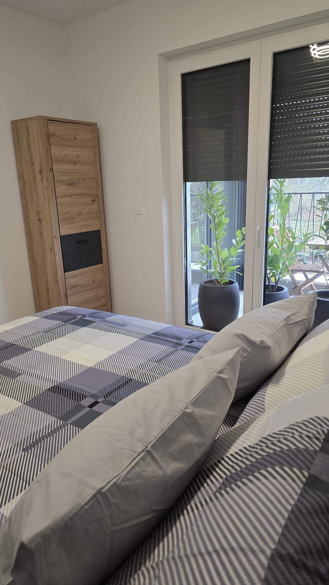 A cozy bedroom is highlighted by a large, comfortable bed adorned with checkered bedding. A wooden storage unit stands nearby, complemented by sliding glass doors that lead to a balcony. Potted plants are visible outside, adding a touch of greenery.