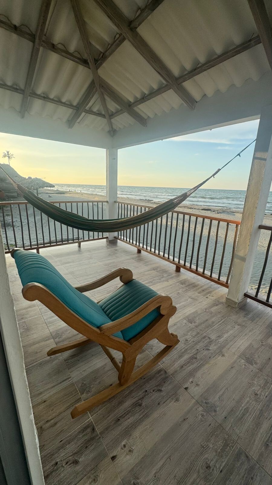 A covered terrace offers a relaxing view of the ocean, featuring a comfortable rocking chair and a hammock suspended nearby. The flooring is modern and wood-like, while the railing provides an open feel to the space, allowing for a seamless connection with the surroundings.