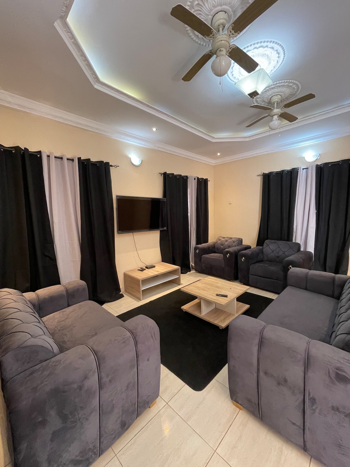A cozy living area features plush seating arranged around a central coffee table. Dark curtains frame the windows, allowing light to contrast with the light-colored walls and tiled floor. A wall-mounted smart TV is positioned for comfortable viewing.