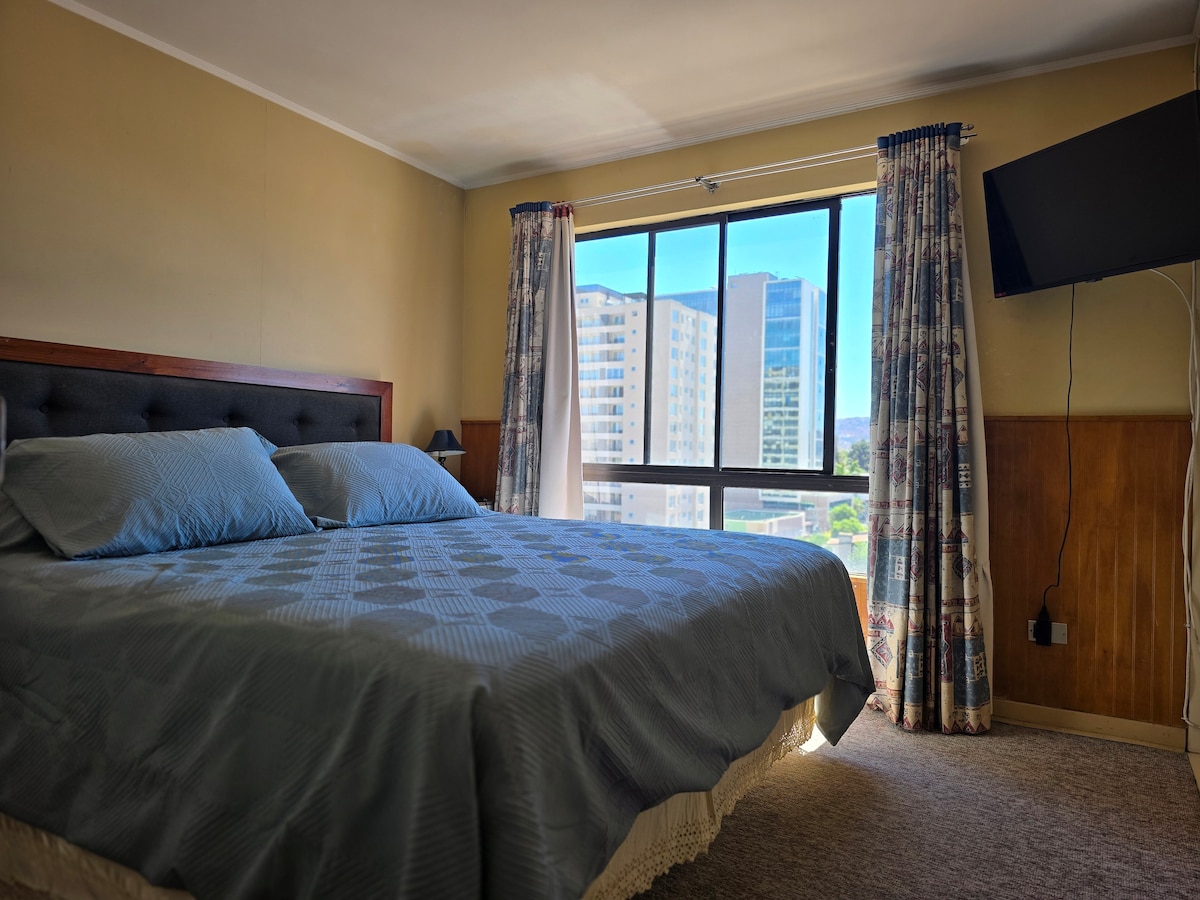 A bright bedroom is equipped with a spacious bed dressed in soft blue bedding. Natural light enters through a large window, revealing views of the cityscape. Soft curtains frame the window, and a flat-screen TV is mounted on the wall opposite the bed.