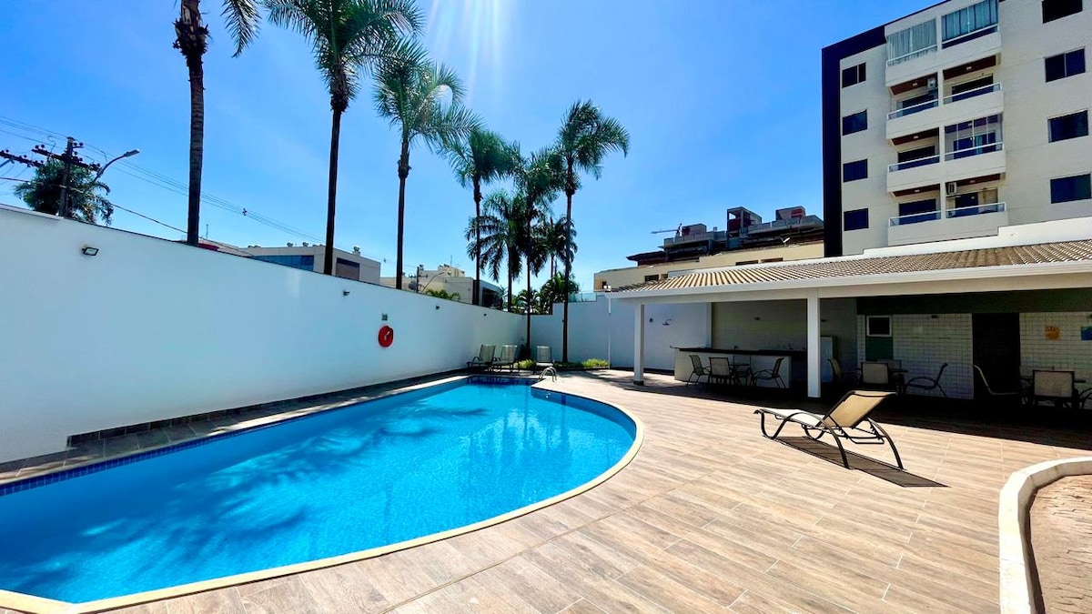 A tranquil outdoor pool is surrounded by palm trees, with lounge chairs positioned neatly on the wooden deck. The clear blue water reflects the sunlight, creating a serene ambiance. The area is enclosed by white walls, providing privacy and a sense of relaxation.