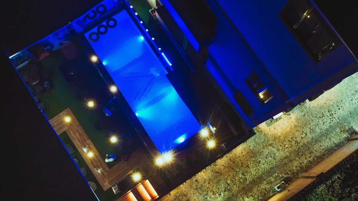 An aerial view highlights a spacious outdoor area featuring a large pool illuminated with soft blue lights. Surrounding spaces include lounges and patios, all complemented by strategically placed lighting that enhances the evening ambiance.