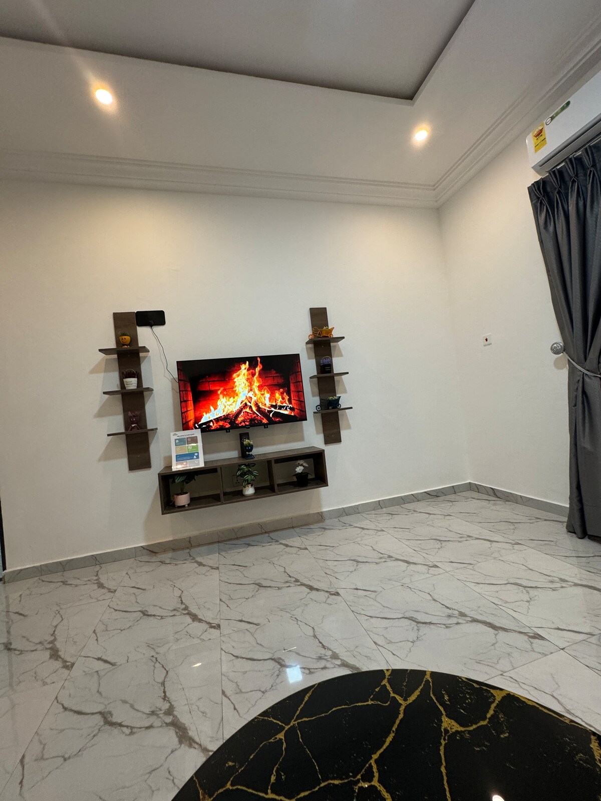 A modern living area features a wall-mounted television displaying a fireplace scene. Shelving units flank the television, providing space for decorative items. A sleek black table with gold accents is positioned in the foreground. The floor is covered with glossy tiles, and soft lighting enhances the ambiance.