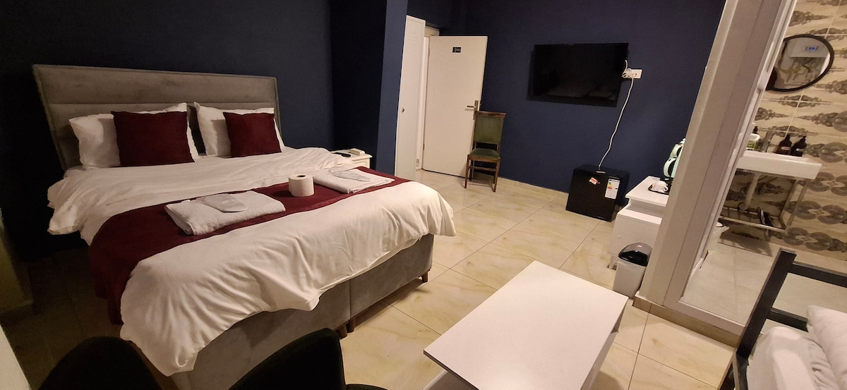 A spacious room features a large bed dressed in white linens with maroon accents. A television is mounted on the wall opposite the bed. A small table and chairs sit nearby, and a modern bathroom area is visible through a glass partition.