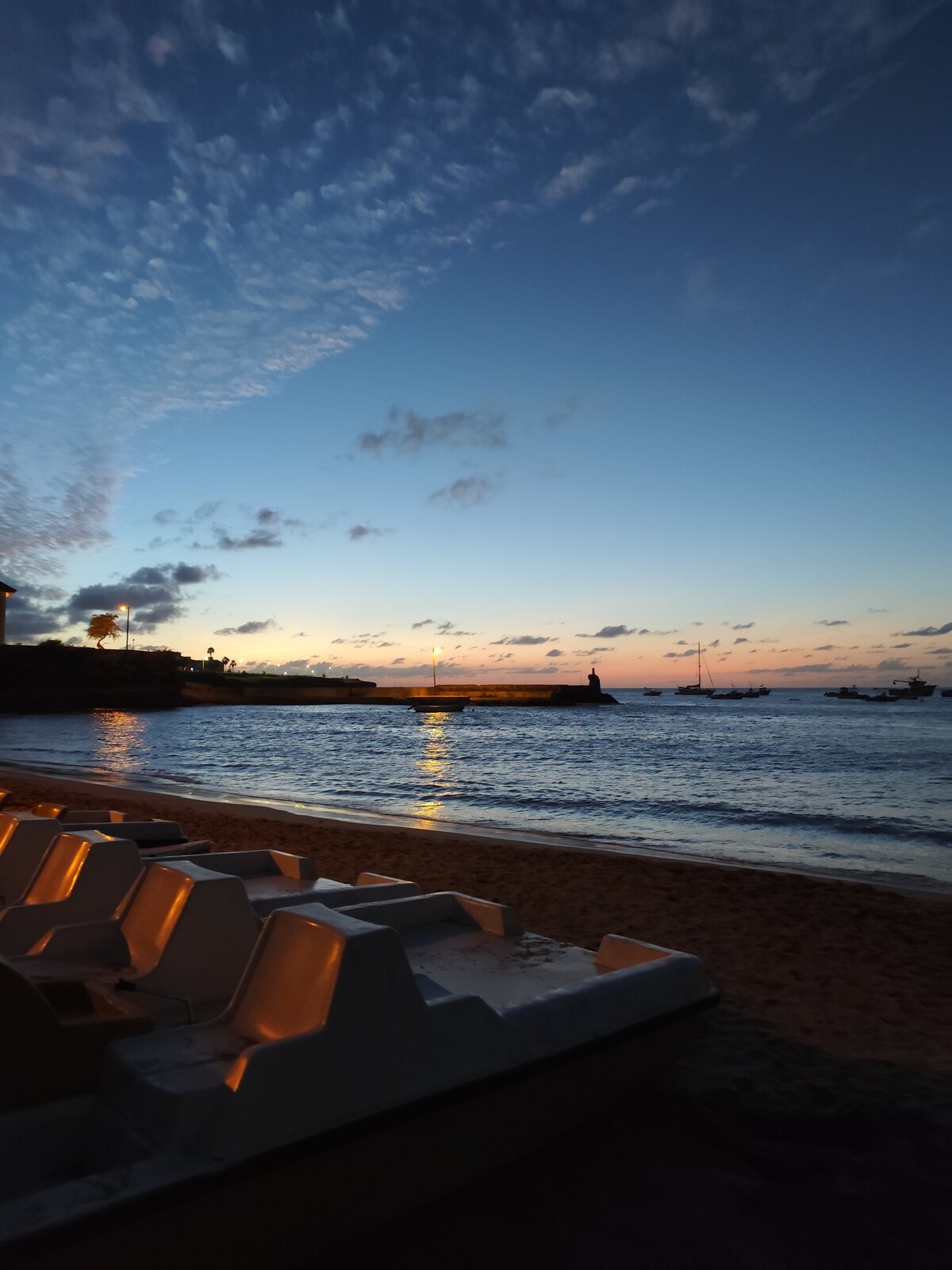 A serene beach scene captures the calm sea reflecting the hues of the sunset. A sandy shoreline is lined with several white lounge chairs, positioned towards the water. Silhouettes of distant boats can be seen against the sky, where scattered clouds are illuminated by a soft golden light.
