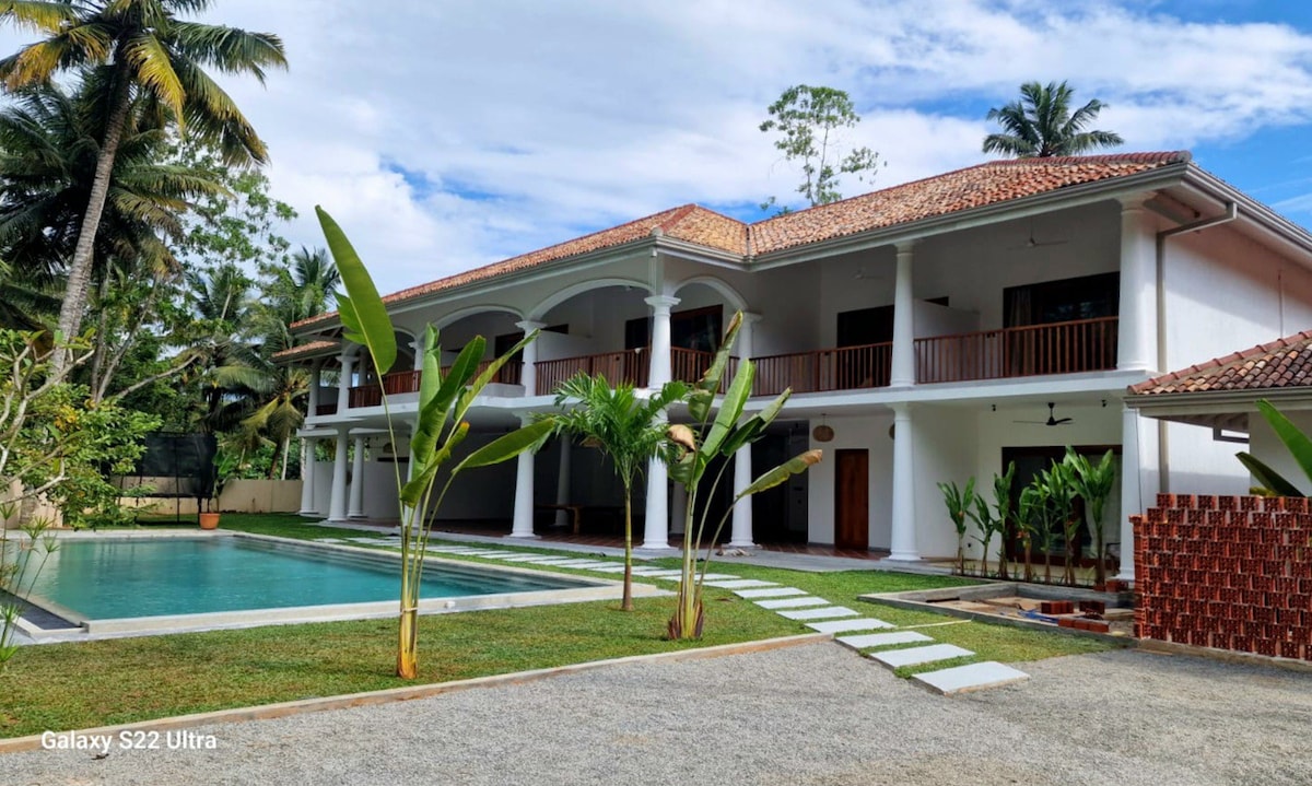 An elegant two-story property is showcased surrounded by lush greenery. A spacious pool is visible in the foreground, complemented by neatly arranged stepping stones leading to the entrance. A few palm trees provide a tropical ambiance against a backdrop of a clear sky.