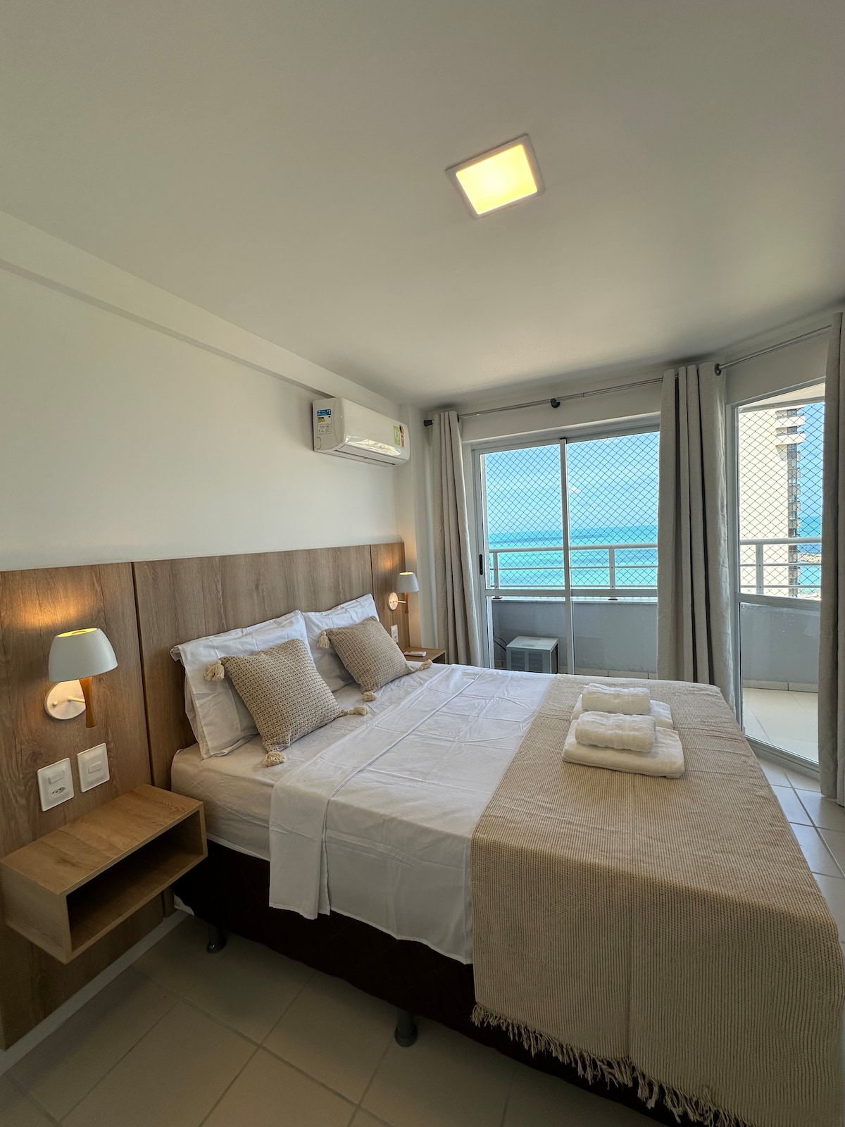 A light-filled bedroom features a spacious bed with neutral-toned bedding. Soft throw blankets are arranged neatly. A bedside table supports reading lights, while a window offers a view of the blue sea beyond. Air conditioning unit is mounted for comfort.