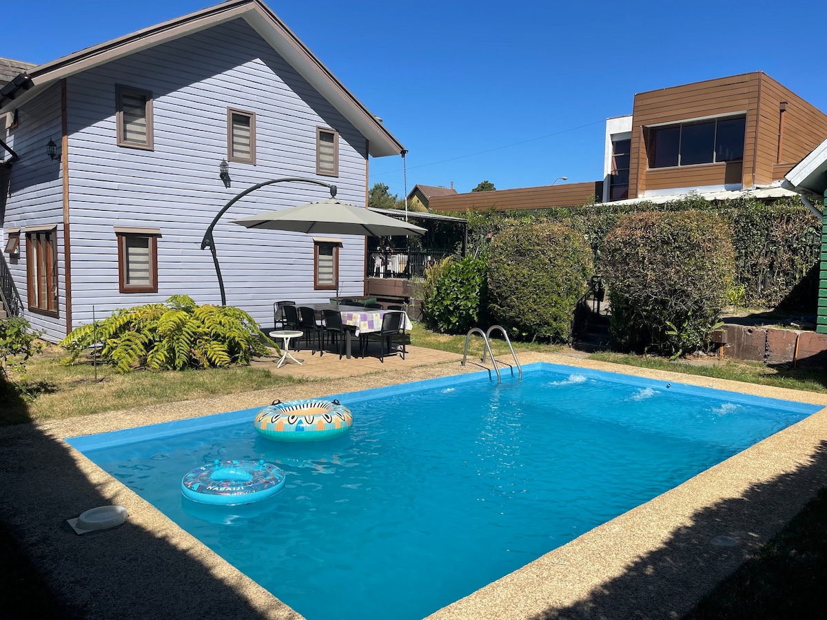 A private swimming pool is surrounded by greenery, featuring a relaxing atmosphere. Lounge chairs and an umbrella are positioned by the poolside. Two inflatable pool floats rest on the water's surface, and a two-story house stands in the background, showcasing a serene outdoor space.
