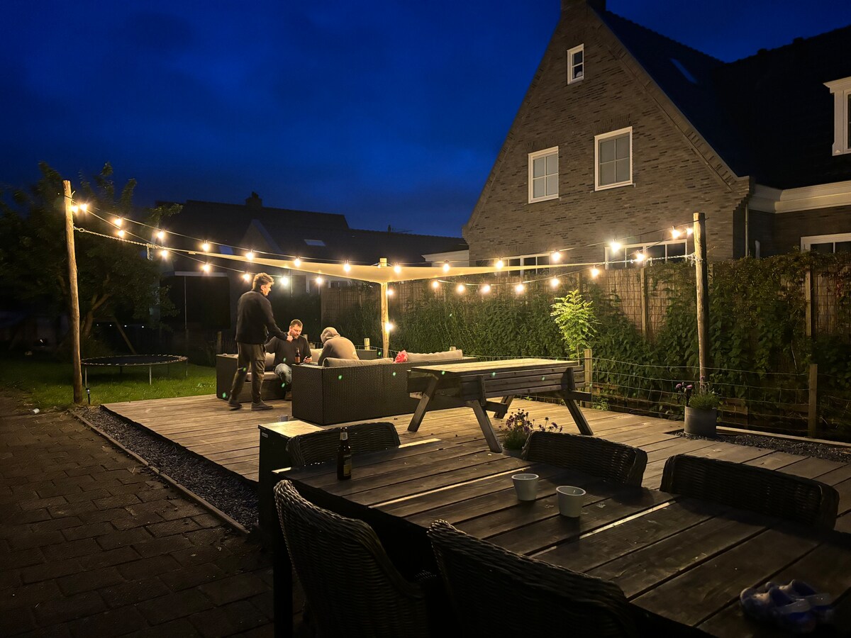 A spacious outdoor area is illuminated by string lights, creating a warm atmosphere. A wooden deck features a seating arrangement with people gathered around a table. A table with a few cups sits nearby, and greenery is visible along the perimeter.