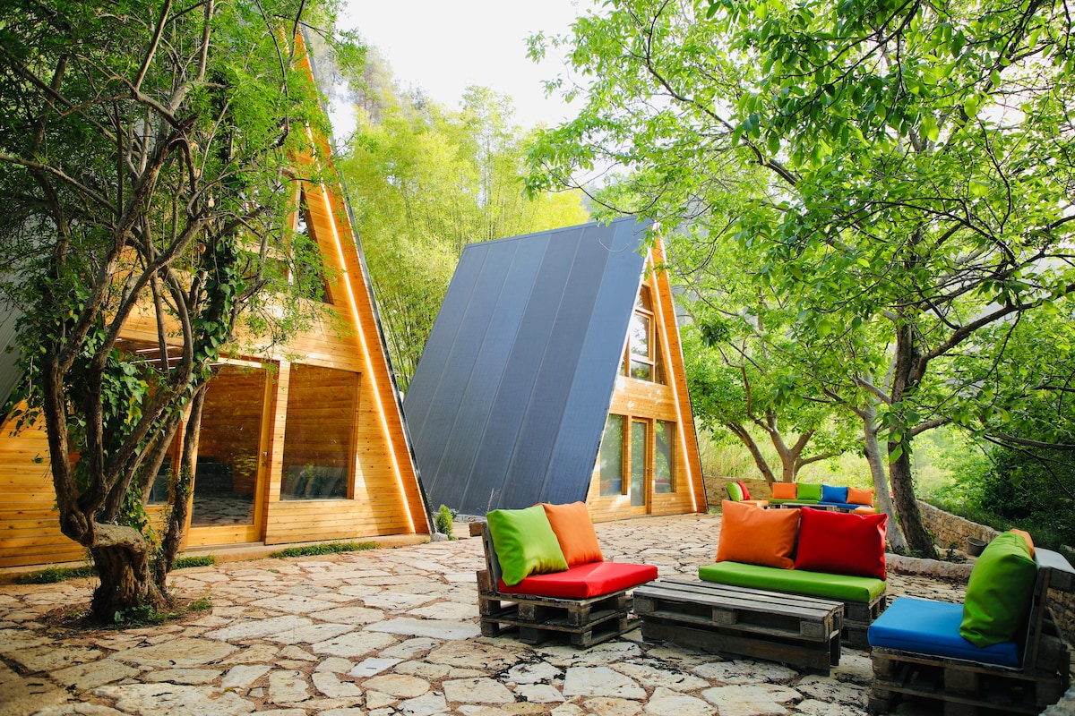 An A-frame cabin is surrounded by lush greenery, featuring a mix of wood and modern elements. Colorful outdoor seating made from repurposed pallets is positioned on a stone patio, inviting relaxation under dappled sunlight filtering through the trees.