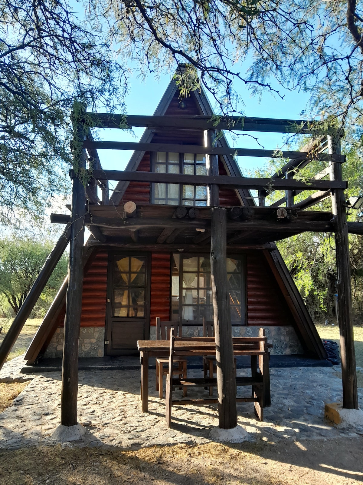 An A-frame cabin is showcased with wooden siding and a stone foundation. A covered porch features a dining table and wooden chairs, surrounded by trees. The entryway is framed by large windows, allowing natural light to illuminate the interior.