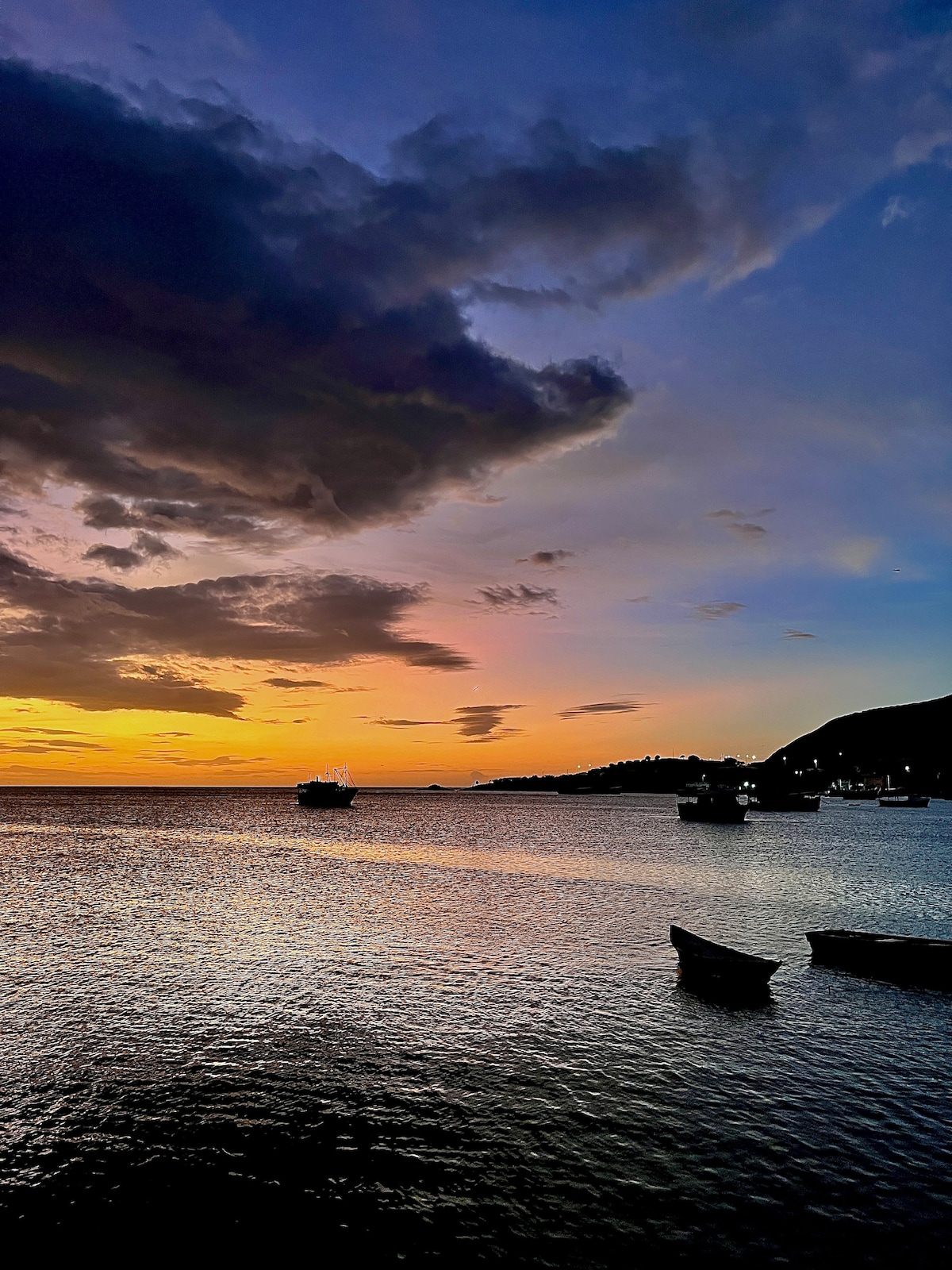 The image captures a calming seascape at sunset, where the sky transitions from deep blues to warm oranges and purples. Silhouetted boats are visible on the water, with gentle ripples reflecting the colorful sky, creating a serene atmosphere.
