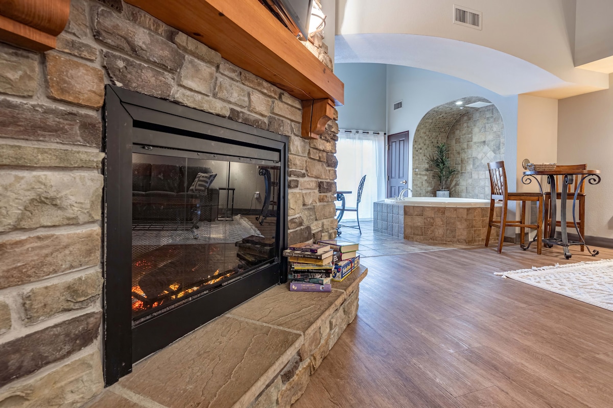 A stone fireplace is positioned next to a wooden mantel, with a stack of books resting on the stone base. In the background, a jacuzzi is visible, along with a dining area featuring high-top chairs. Natural light enters from a nearby patio door.