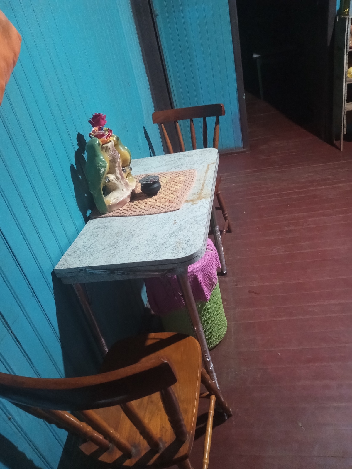 A small dining area features a rustic table with a textured surface, accompanied by two lightly stained wooden chairs. A decorative item and a small cup are placed on a woven mat atop the table, set against a bright blue wall.