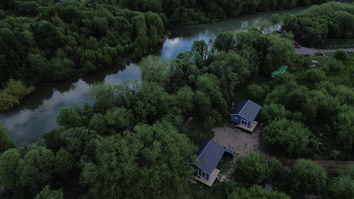 An aerial view displays a serene landscape featuring two cottages surrounded by lush greenery. A calm river meanders nearby, reflecting the surrounding trees. The natural setting creates a peaceful atmosphere, ideal for families seeking relaxation.