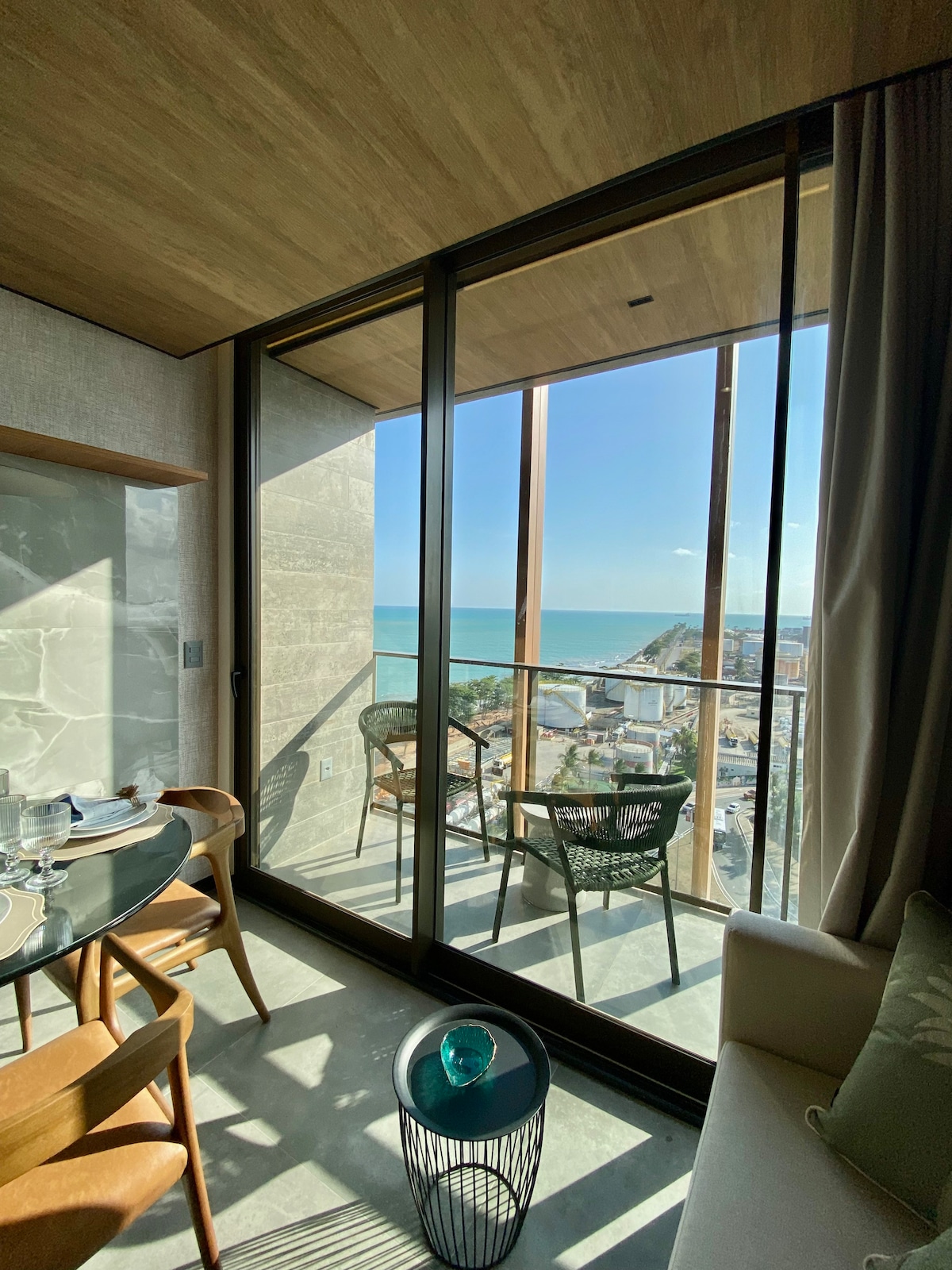 A bright seating area features a round dining table with elegant chairs. Floor-to-ceiling windows provide a panoramic view of the sea and coastline. The serene space is bathed in natural light, creating an inviting atmosphere while highlighting a tastefully decorated balcony.