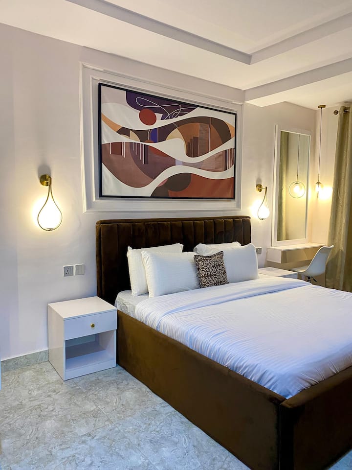 Пространа спална соба со брачен кревет, придружена со простор наменет за работа кој се дуплира како простор за облекување, опремена со огледало и две светилки, додека креветот е опкружен со две дополнителни светилки.
