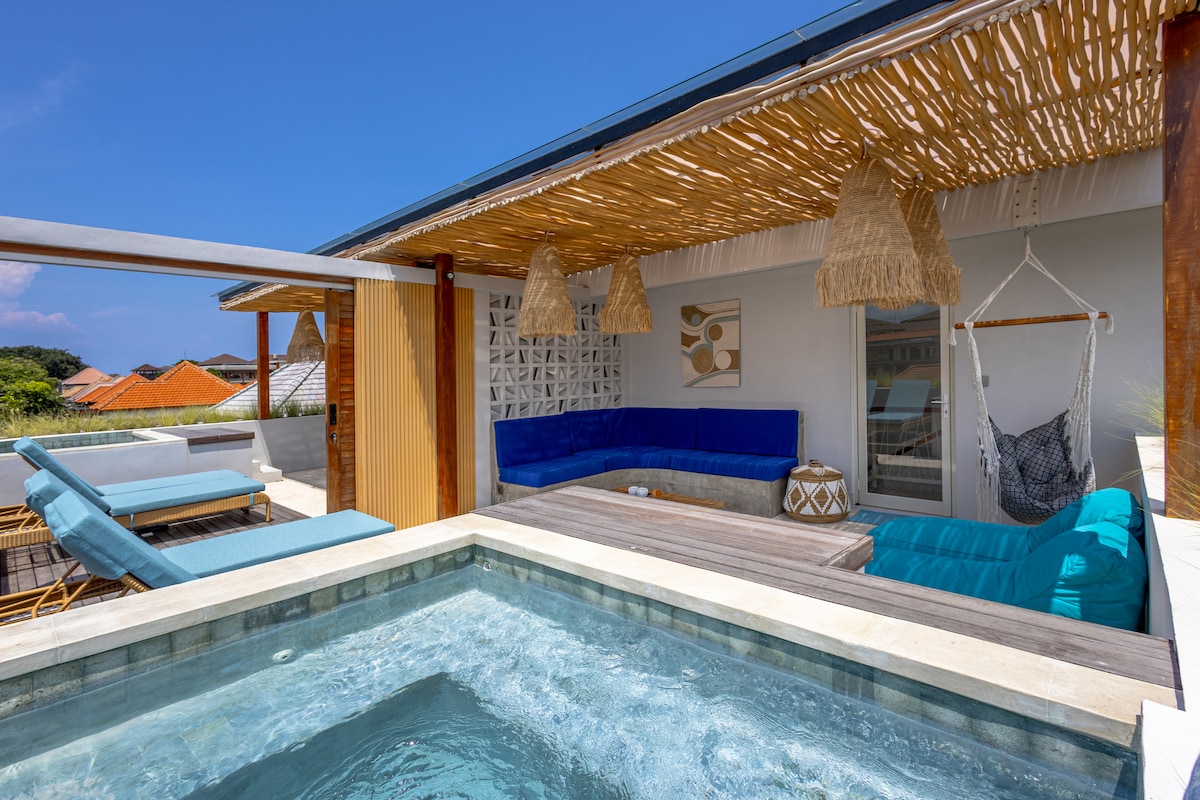 A rooftop terrace features a plunge pool surrounded by a wooden deck. Comfortable seating areas, including a blue sectional sofa and a hanging chair, are arranged under a thatched roof. Bright blue skies and scenic views are visible in the background.